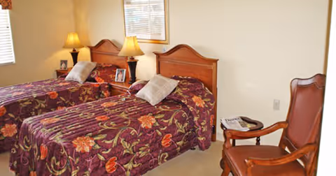 A bedroom with two single beds featuring wooden headboards and floral patterned bedspreads in shades of purple, red, and green. Each bed has a pillow and a bedside table with a lamp and framed photos. There is a wooden chair with armrests next to the beds, and a window with blinds letting in natural light.