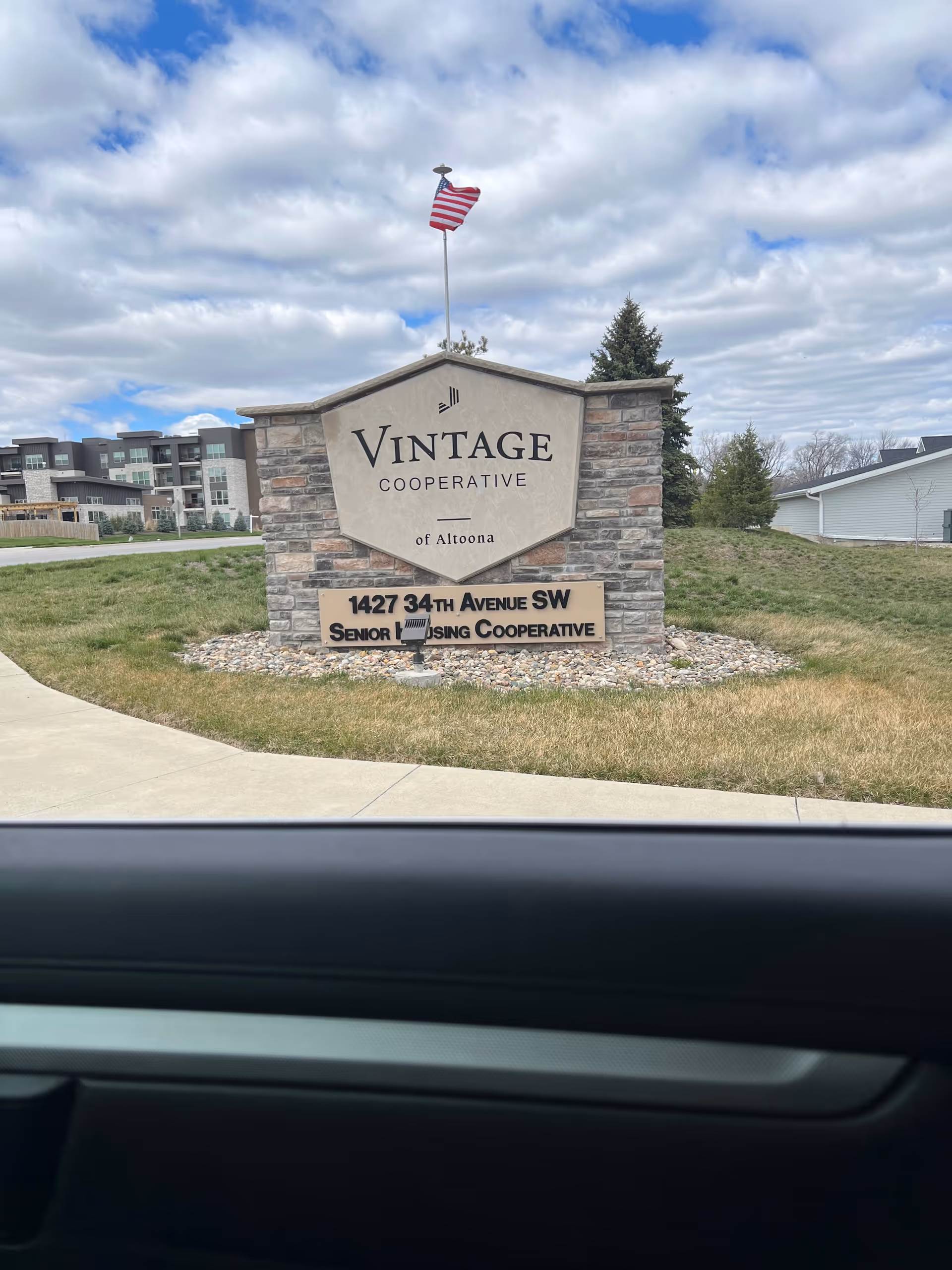Stone sign for Vintage Cooperative of Altoona with an American flag on top, located on a grassy area next to a sidewalk. Residential buildings and a house are visible in the background under a partly cloudy sky.