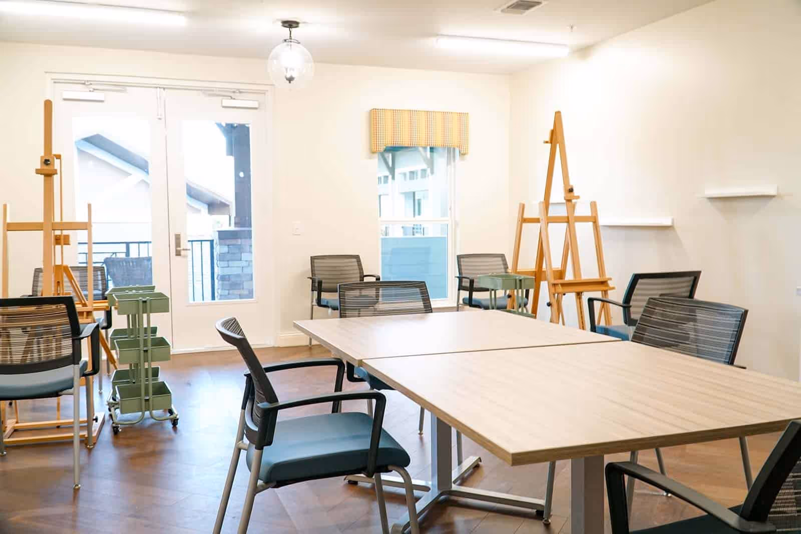 A bright, spacious room with several chairs around two rectangular tables pushed together, wooden easels, and a green rolling cart. The room has large windows and a glass door leading outside, with light-colored walls and wood flooring.