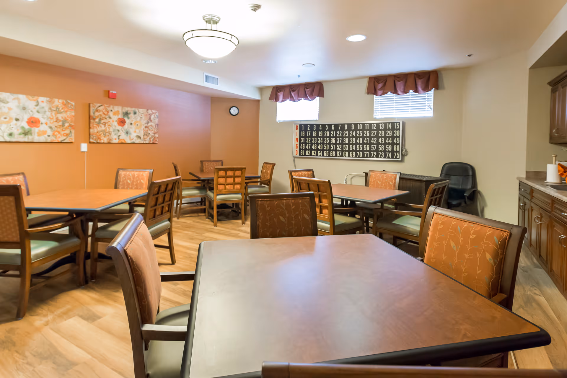 Dining/activity room with multiple tables and chairs, a bingo board on the wall, and warm decor.