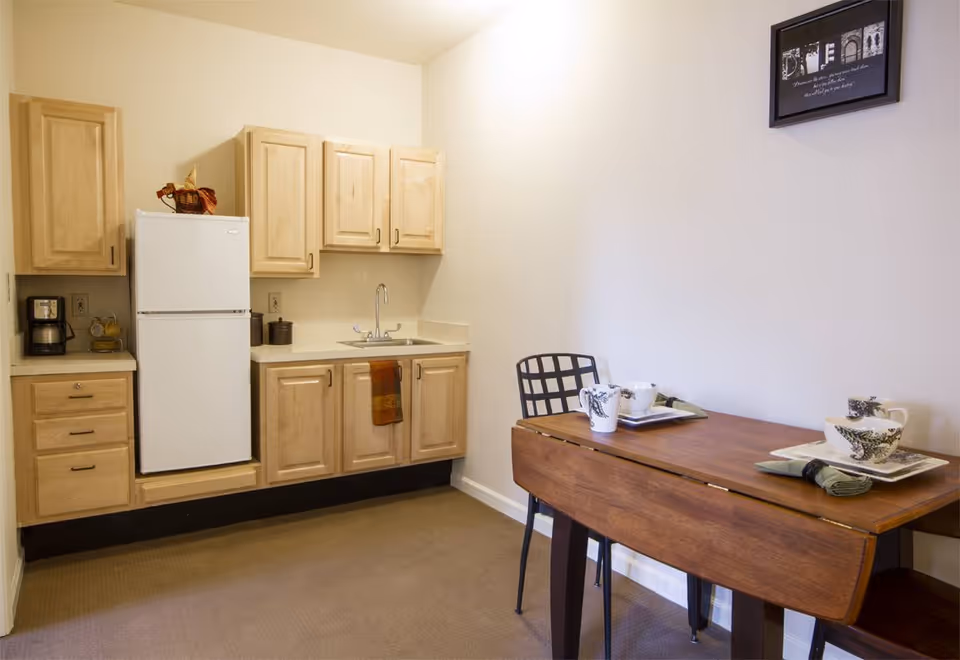 A small kitchen area with light wood cabinets, a white refrigerator, a coffee maker, and a sink with a towel hanging from the cabinet handle. To the right, there is a wooden drop-leaf table set with two place settings including cups, plates, and napkins, accompanied by a black metal chair. A framed picture hangs on the wall above the table.