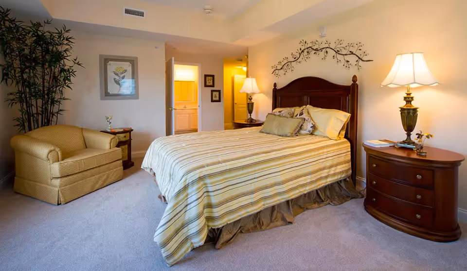 Well-lit bedroom with a striped bed and wooden headboard, bedside tables with lamps, an armchair, plant, and a doorway leading to a bathroom.