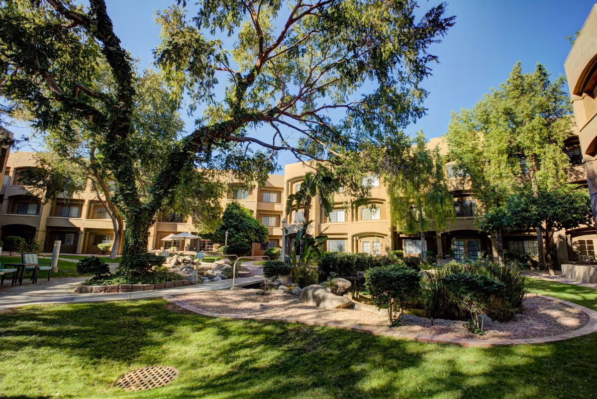 Outdoor courtyard area of a senior living facility with large trees, green grass, shrubs, and a rock garden. Surrounding the courtyard are multi-story beige buildings with windows and balconies under a clear blue sky.