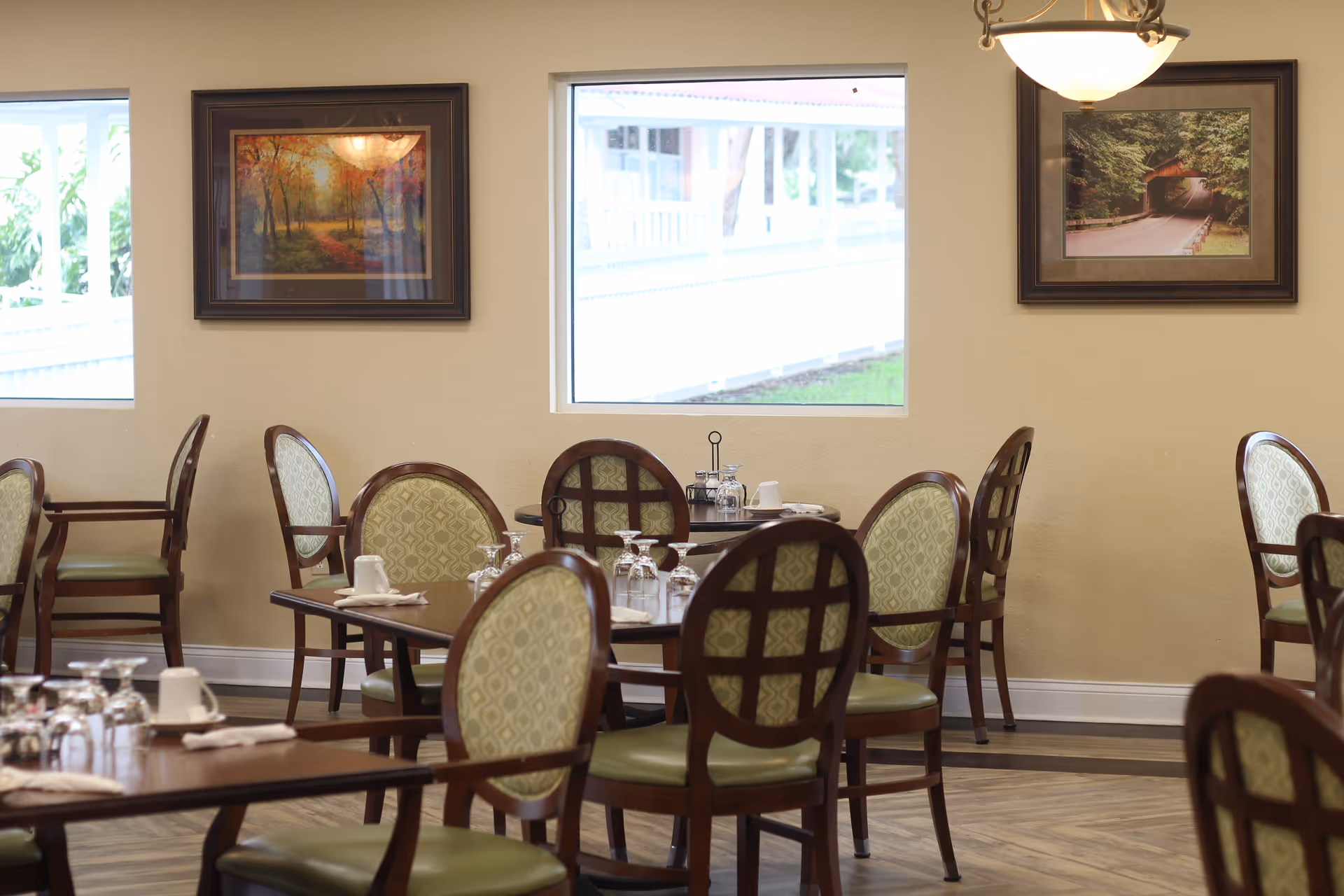 Dining area with wooden tables and chairs featuring green patterned upholstery. The tables are set with upside-down glasses, white cups, and napkins. Two framed landscape paintings hang on the beige walls, and two windows provide natural light with a view of the outside. A ceiling light fixture is also visible.