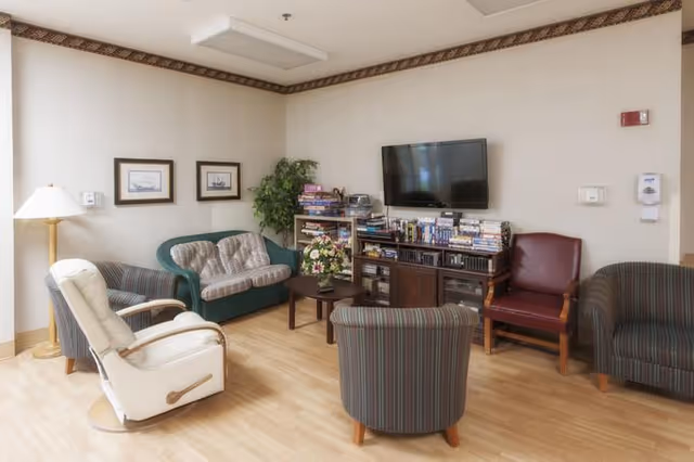 A cozy living room area with a variety of seating including a white recliner, a green loveseat, a red armchair, and two striped chairs arranged around a small wooden coffee table with a flower arrangement. A wooden entertainment center holds a flat-screen TV and several stacks of books and DVDs. The walls are decorated with two framed pictures and a potted plant is in the corner. The room has light wood flooring and a ceiling light fixture.
