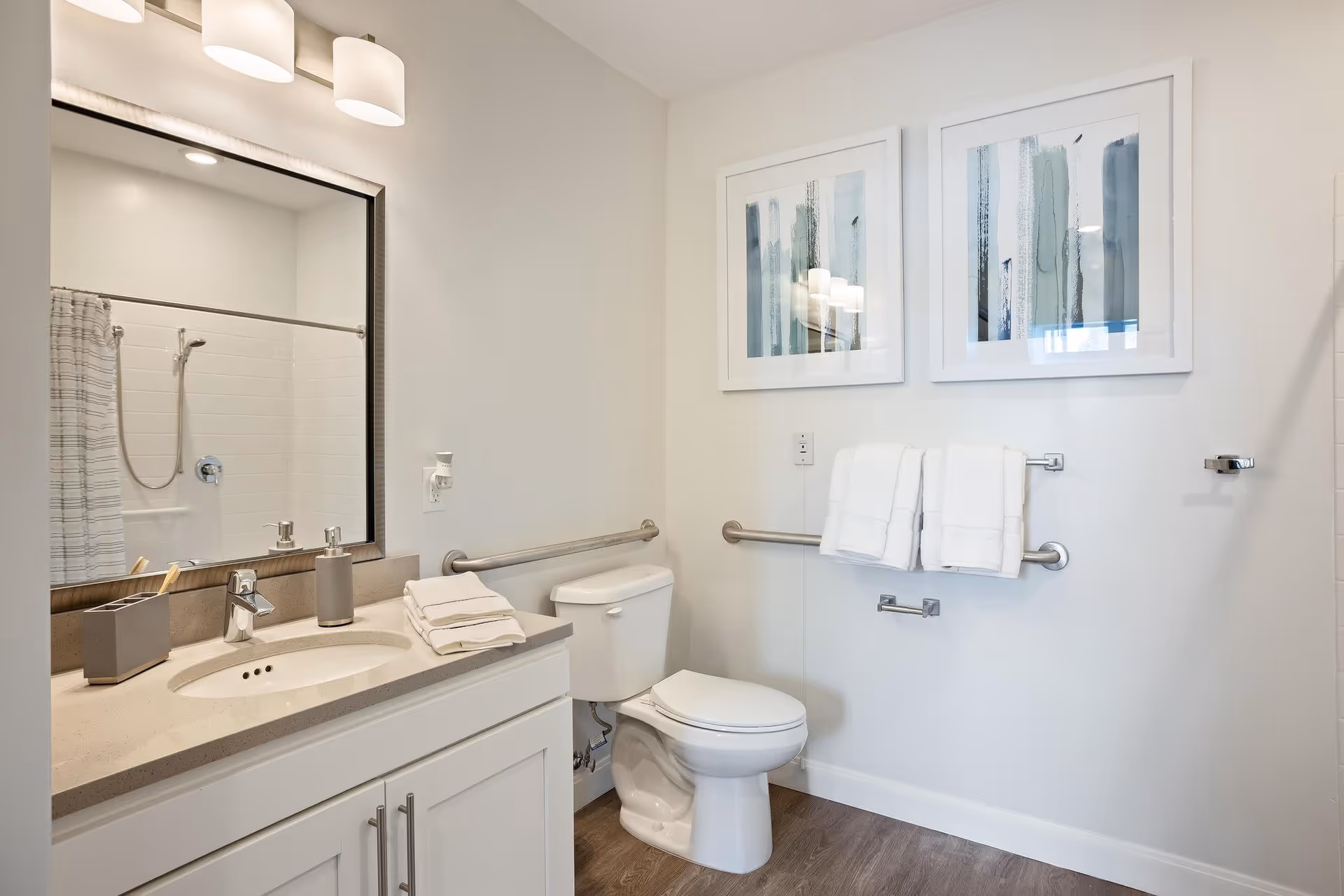 A clean and modern bathroom featuring a white vanity with a sink, a large mirror with two light fixtures above it, a toilet with grab bars on the walls for accessibility, two framed abstract paintings on the wall, and white towels hanging on a towel rack.