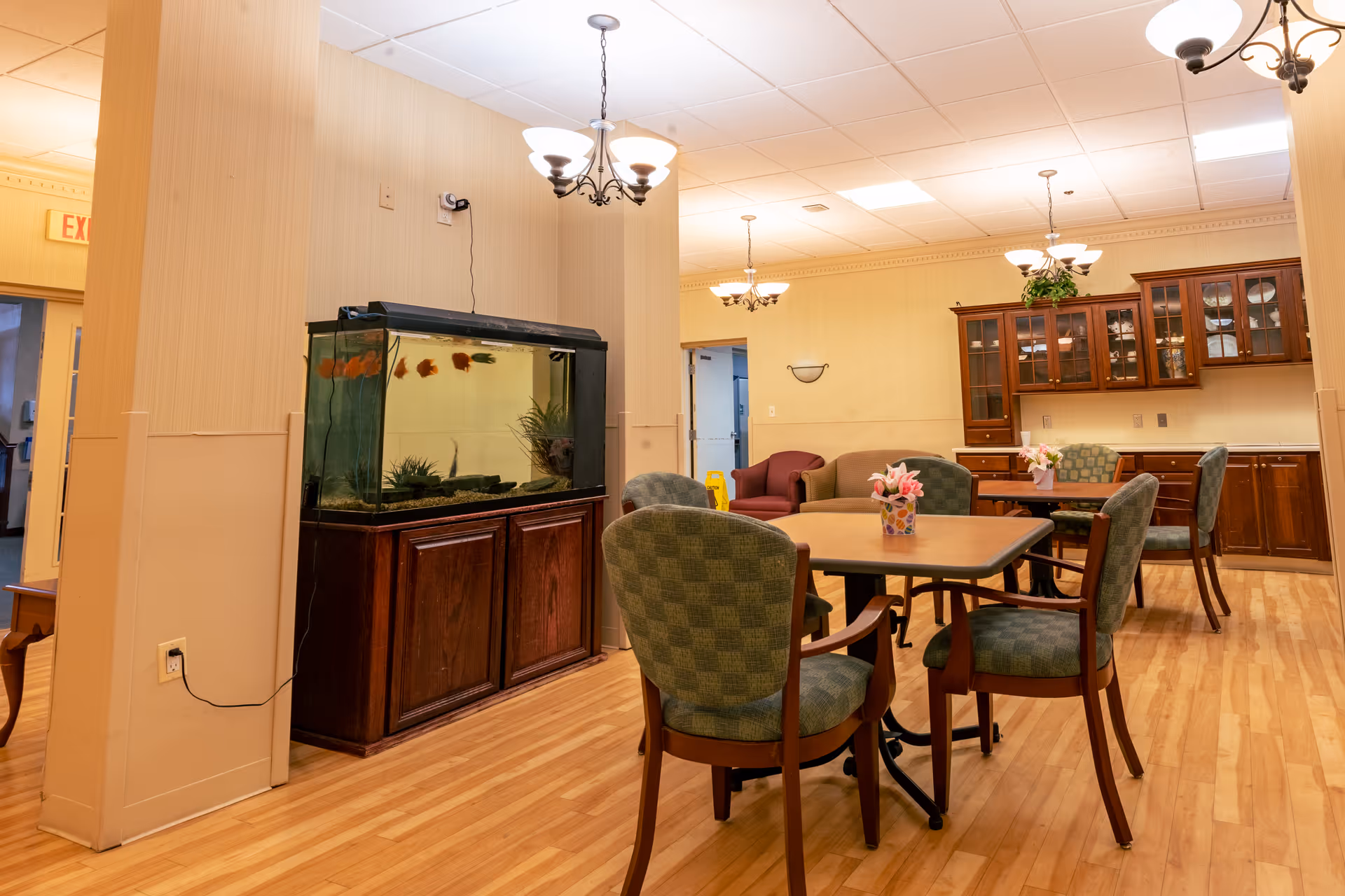 A cozy common area with wooden flooring, several tables and green cushioned chairs, a large fish tank on a wooden cabinet, and a wooden cabinet with glass doors in the background. The room is softly lit by ceiling lights and chandeliers, with a small flower vase on each table.