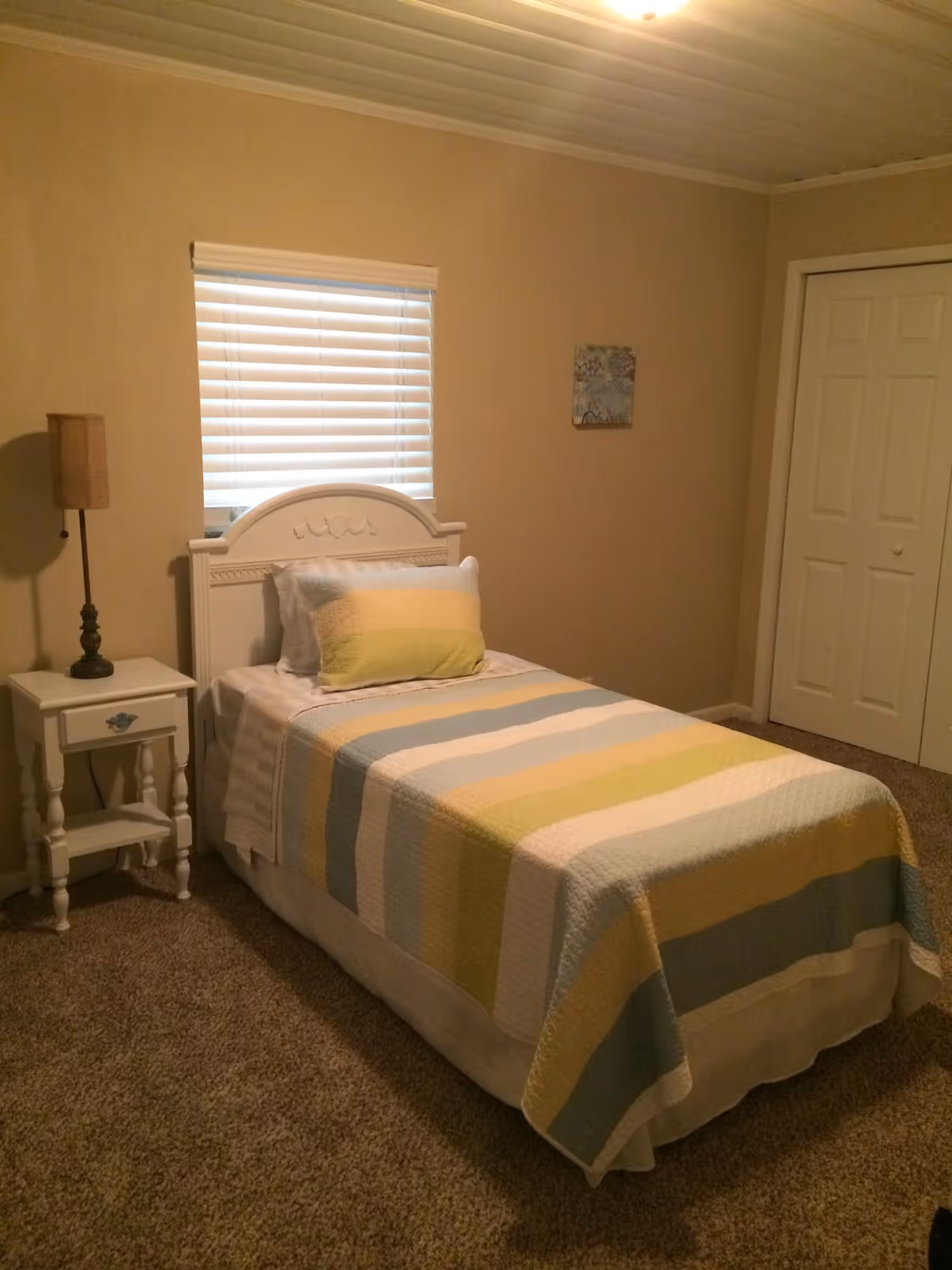 A small bedroom with a single bed covered in a striped quilt with pastel colors including yellow, green, blue, and white. There is a white headboard, a matching white nightstand with a lamp, a window with closed blinds, and a closed closet door. The walls are beige and the floor is carpeted.