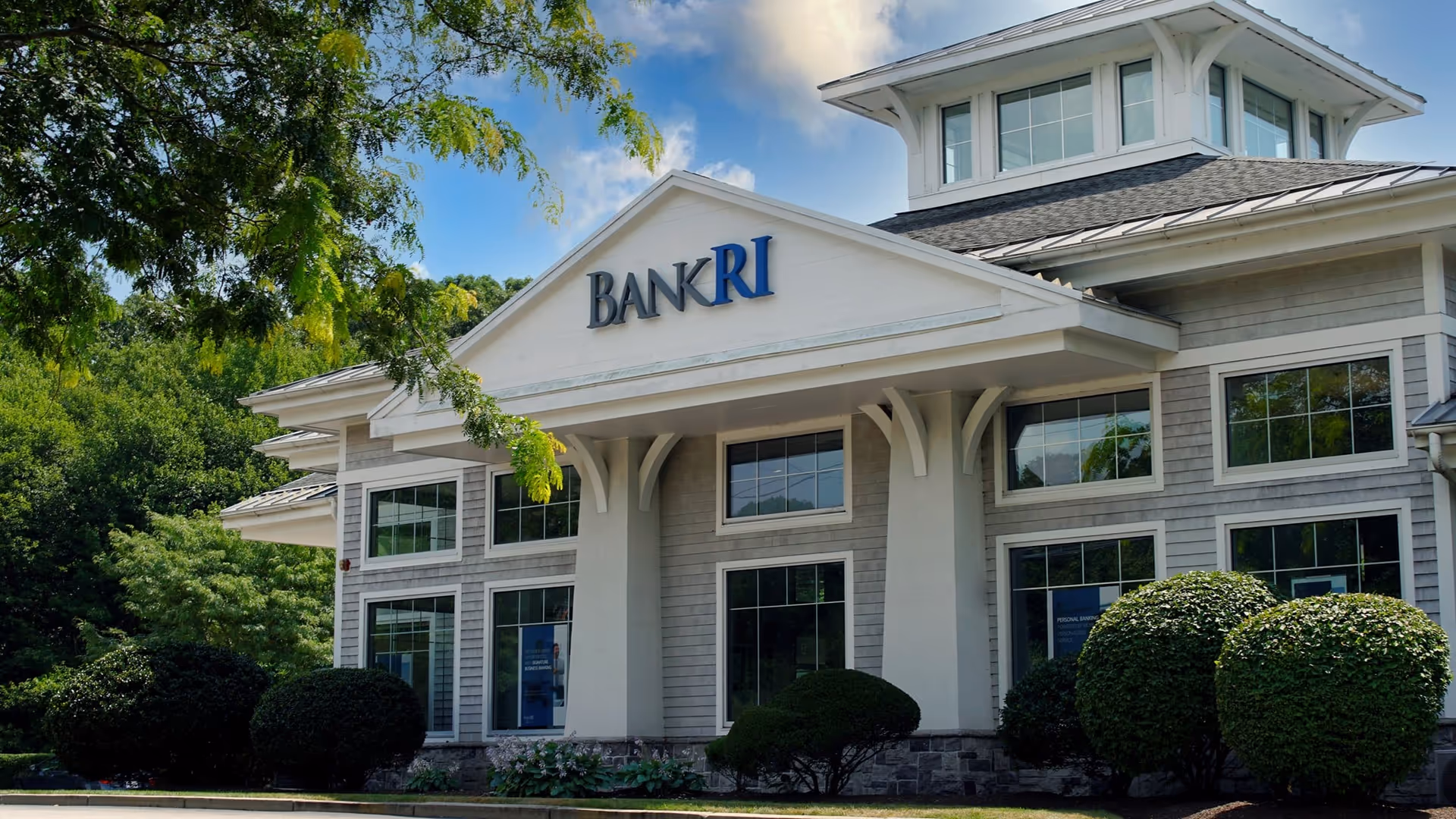 Exterior view of a BankRI building with large windows, a peaked roof, and surrounding green bushes and trees under a partly cloudy blue sky.
