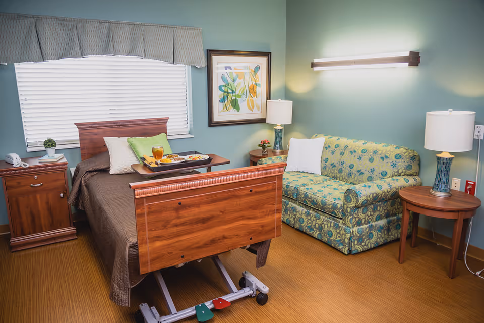A well-lit room with a hospital-style adjustable wooden bed covered with a brown blanket and pillows, a tray with food and a drink on the bed, a wooden nightstand with a phone and a small plant, a patterned green and blue couch with a white pillow, two side tables with lamps, a framed abstract artwork on the wall, and a window with blinds and a valance.