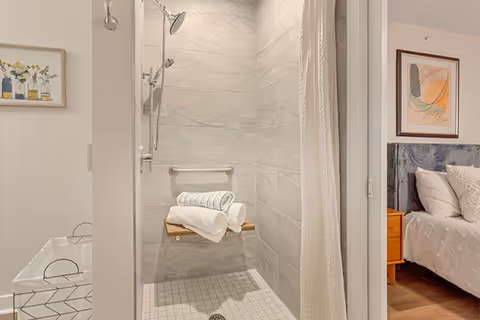 View of a bathroom shower area with light gray tiled walls and floor, a mounted showerhead, and a foldable wooden shower seat holding neatly folded white towels. To the right, a glimpse of a bedroom with a bed, pillows, a wooden nightstand, and framed artwork on the wall is visible.