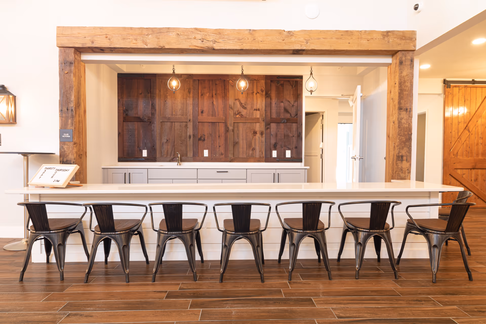 A modern kitchen area with a long white countertop and eight black metal chairs lined up in front. The back wall features dark wooden cabinets and three hanging pendant lights. The space has wooden beams and flooring, with a barn-style sliding door visible on the right.