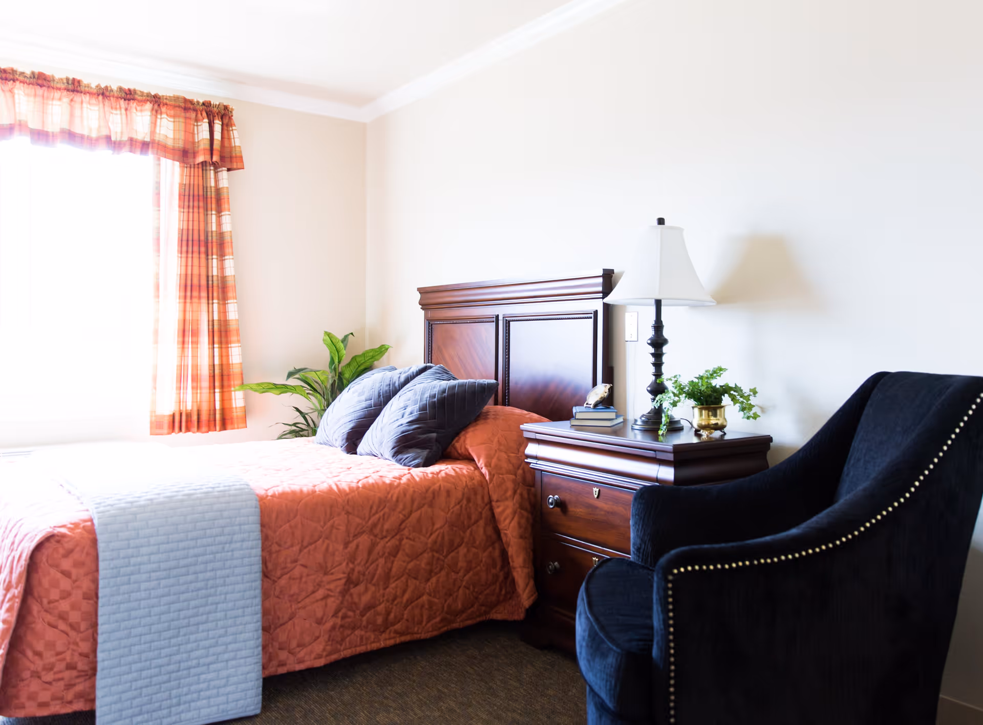A cozy bedroom with a single bed covered in an orange quilt and a light blue throw. The bed has two dark blue pillows and a wooden headboard. Next to the bed is a wooden nightstand with a white lamp, a small plant, and a few books. A dark blue upholstered armchair with nailhead trim is positioned beside the nightstand. A window with orange plaid curtains allows natural light to brighten the room.