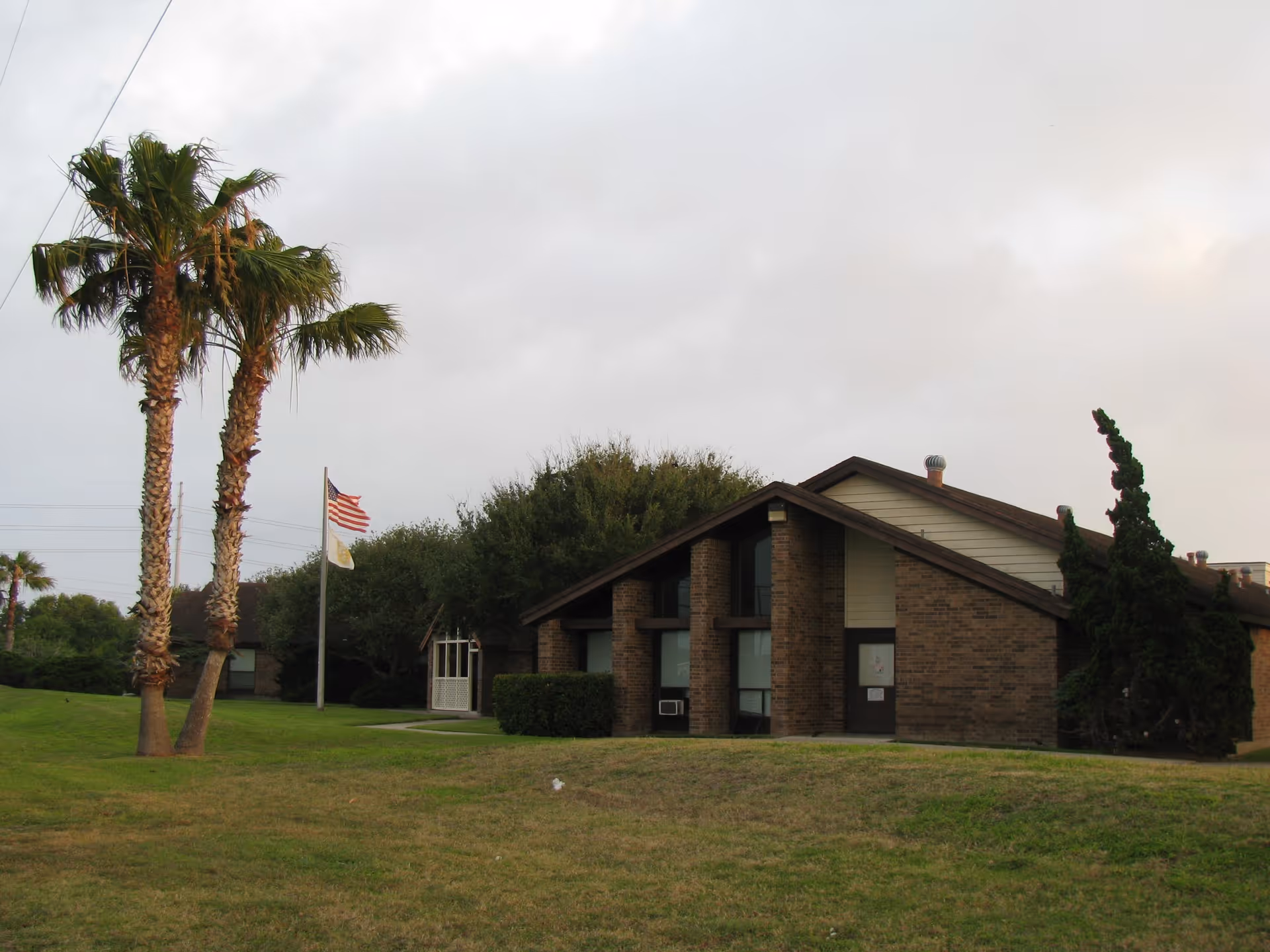 Exterior view of a single-story brick building with a sloped roof, surrounded by green grass and trees including palm trees. An American flag and another flag are flying on a flagpole near the building under a cloudy sky.