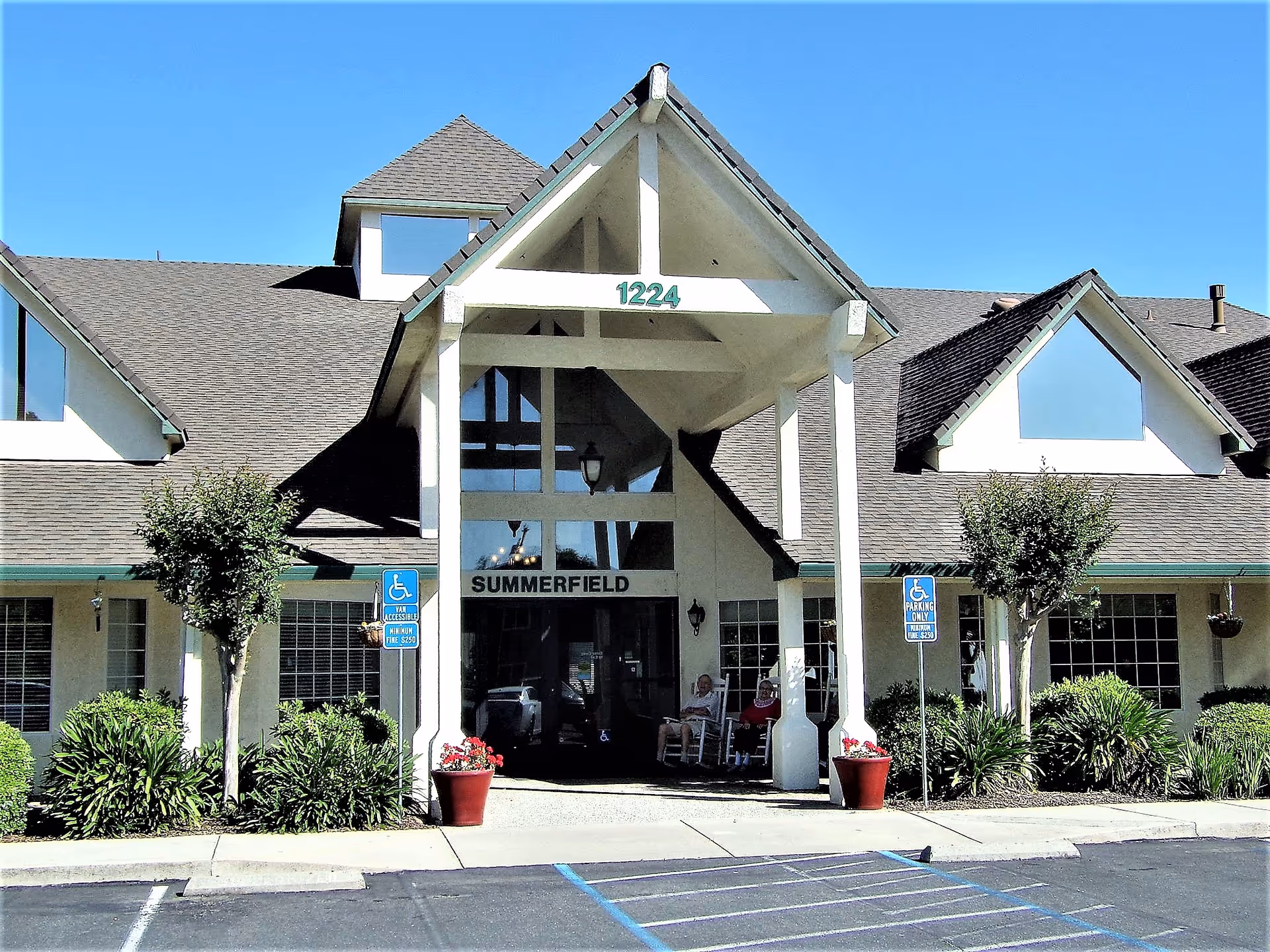 Front exterior view of Summerfield Senior Living facility with a peaked roof entrance, two people sitting on rocking chairs under the covered porch, and handicap parking spaces in front.