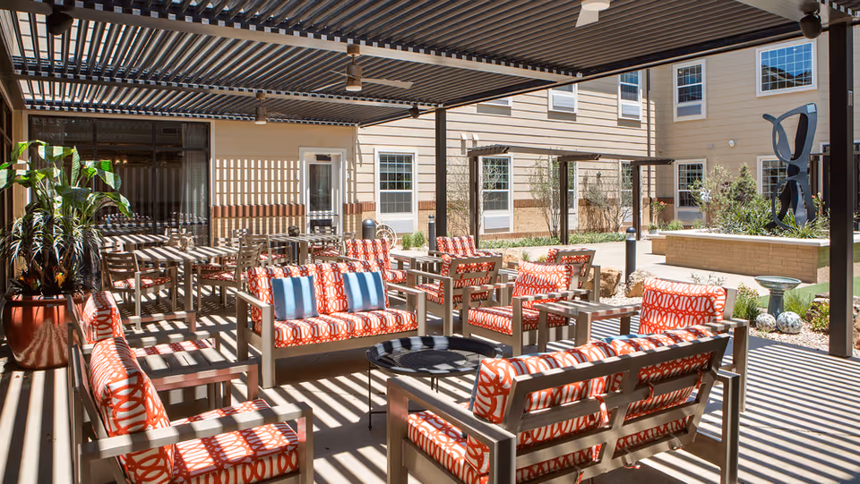 Shaded outdoor courtyard with patterned red-cushioned chairs and tables under a pergola beside a senior living building.
