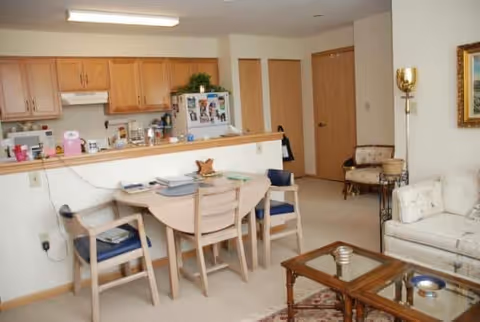 Interior view of a senior living facility apartment showing a small kitchen with wooden cabinets and a refrigerator, a dining table with four chairs, and a living area with a sofa, an armchair, a glass coffee table, and a floor lamp. There is a door in the background and some decorative items on the walls and surfaces.
