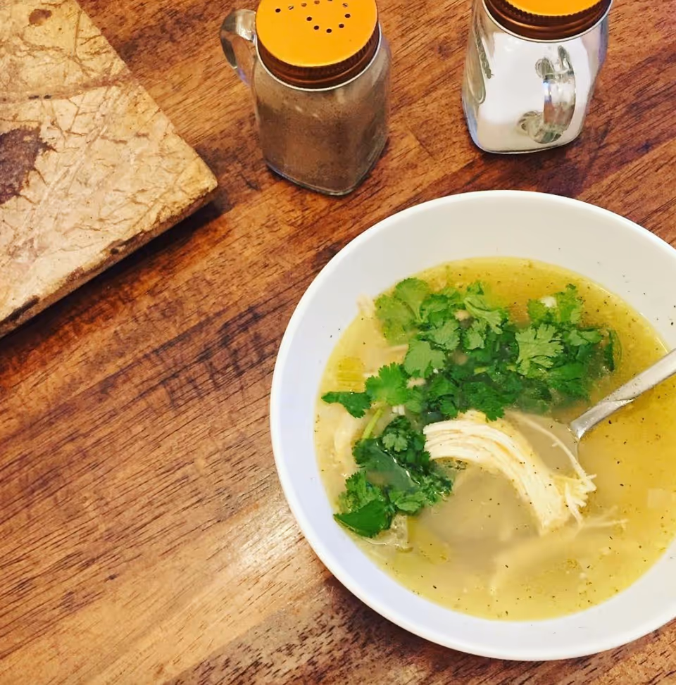 Bowl of chicken soup garnished with cilantro and a spoon on a wooden table beside salt and pepper shakers.