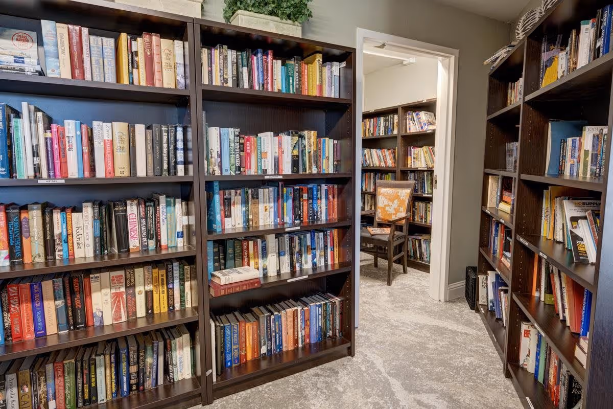 Interior view of a library room with multiple dark wooden bookshelves filled with books. There is a doorway leading to another room with more bookshelves and a wooden chair with an orange and white patterned cushion. The floor is carpeted in a light gray color.
