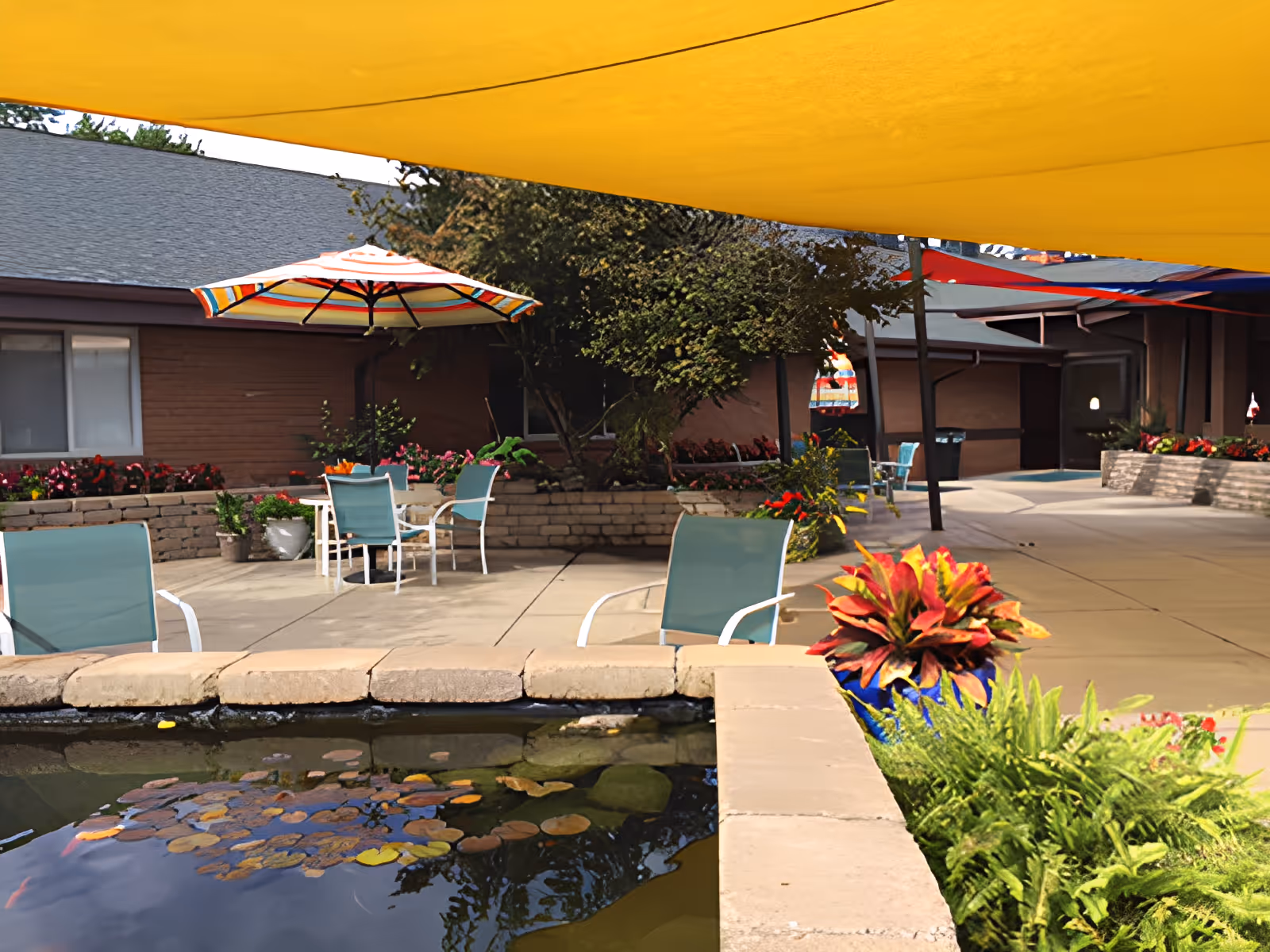 Outdoor patio area at Friends Care Assisted Living with a small pond in the foreground, surrounded by stone edging and plants. There are several chairs and tables with colorful umbrellas providing shade. The area is paved and adjacent to a building with windows and flower beds.