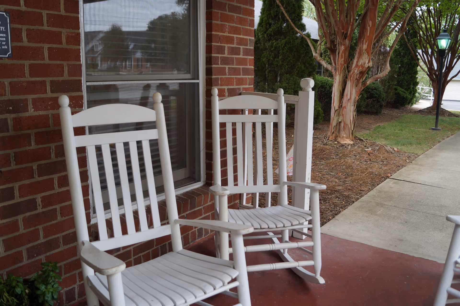 Two white wooden rocking chairs on a red concrete porch next to a brick wall with a window. There is a walkway and some greenery including trees and bushes beside the porch.