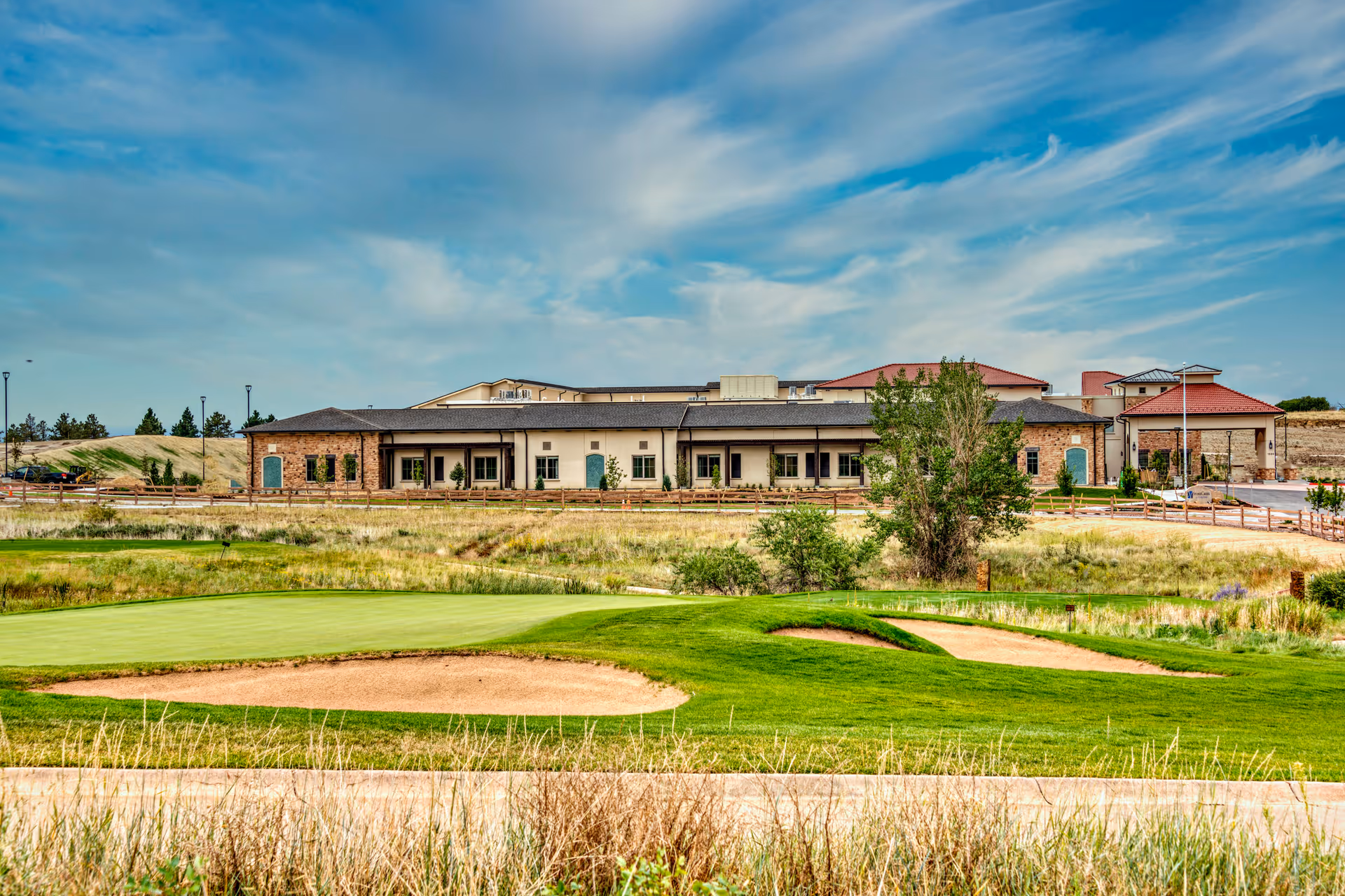 Exterior view of a low-profile senior living building with a manicured golf green and sand bunkers in the foreground under a partly cloudy sky.