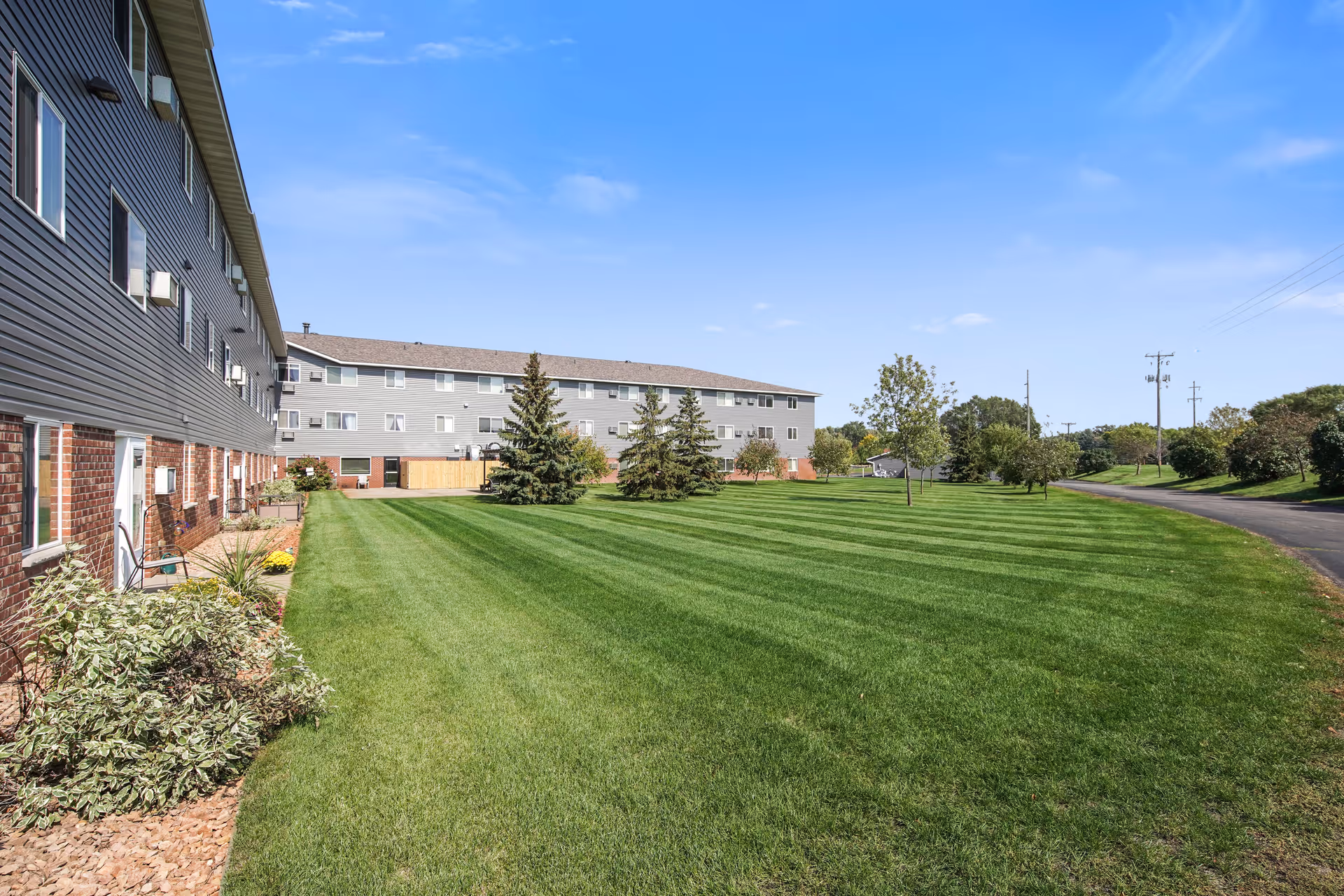 A large, well-maintained grassy lawn with a few trees and shrubs, bordered by a multi-story residential building on the left and a paved road on the right under a clear blue sky.