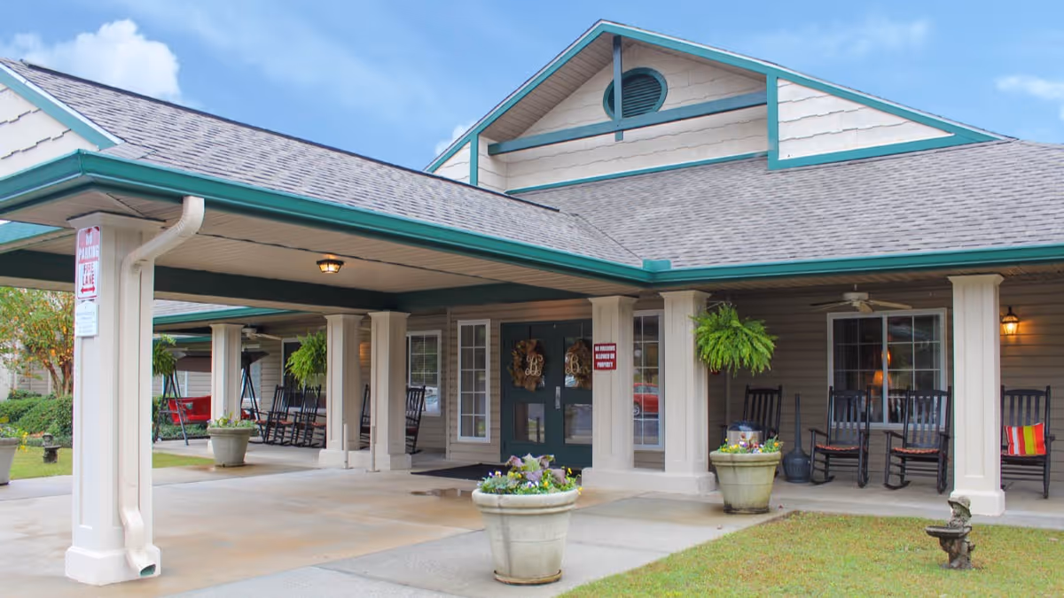 Entrance of a senior living facility with a covered driveway, beige exterior walls, green trim, several rocking chairs on the porch, hanging plants, and potted flowers near the entrance.