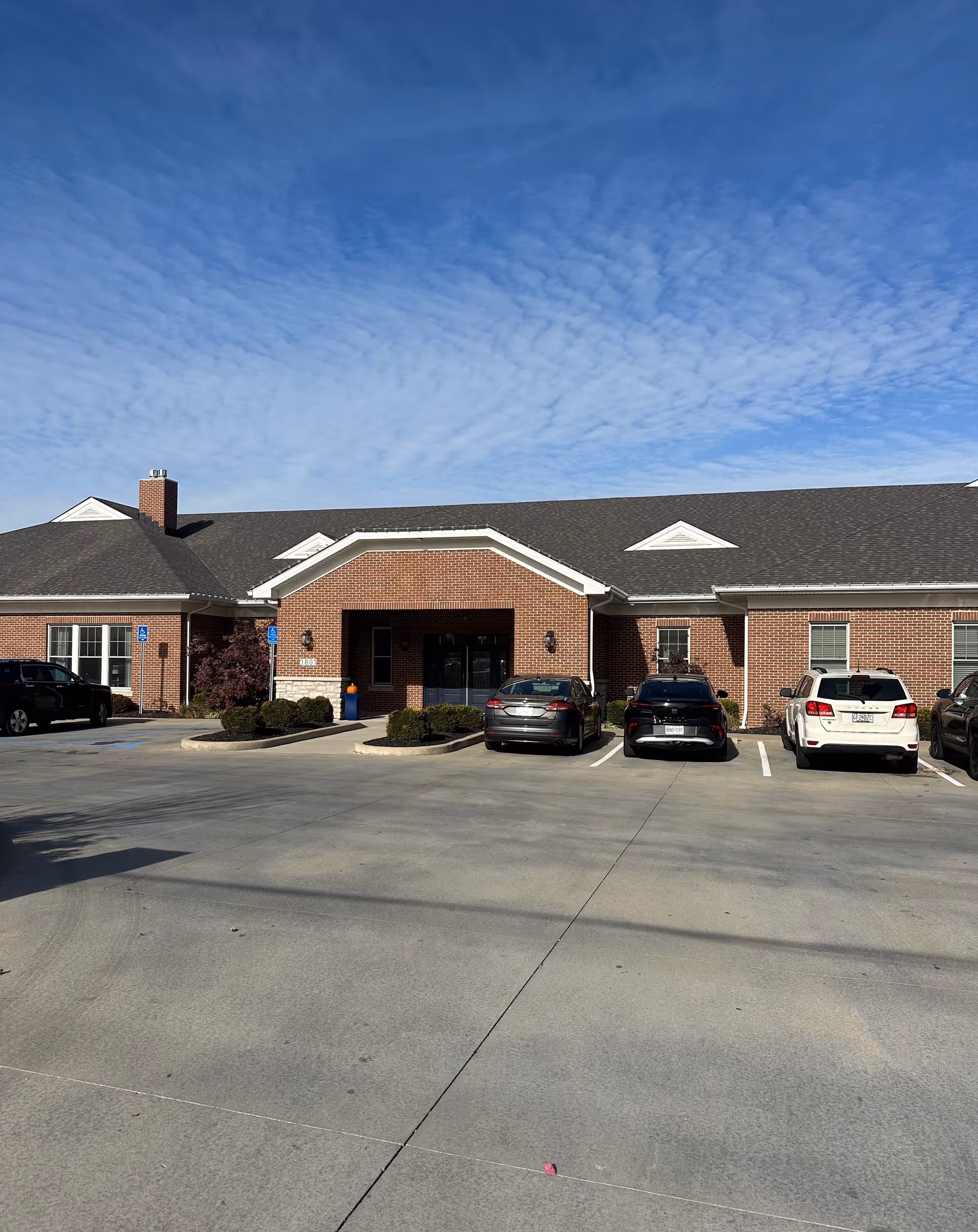 Front exterior of a single-story brick senior living building with a parking lot and several cars under a blue sky.