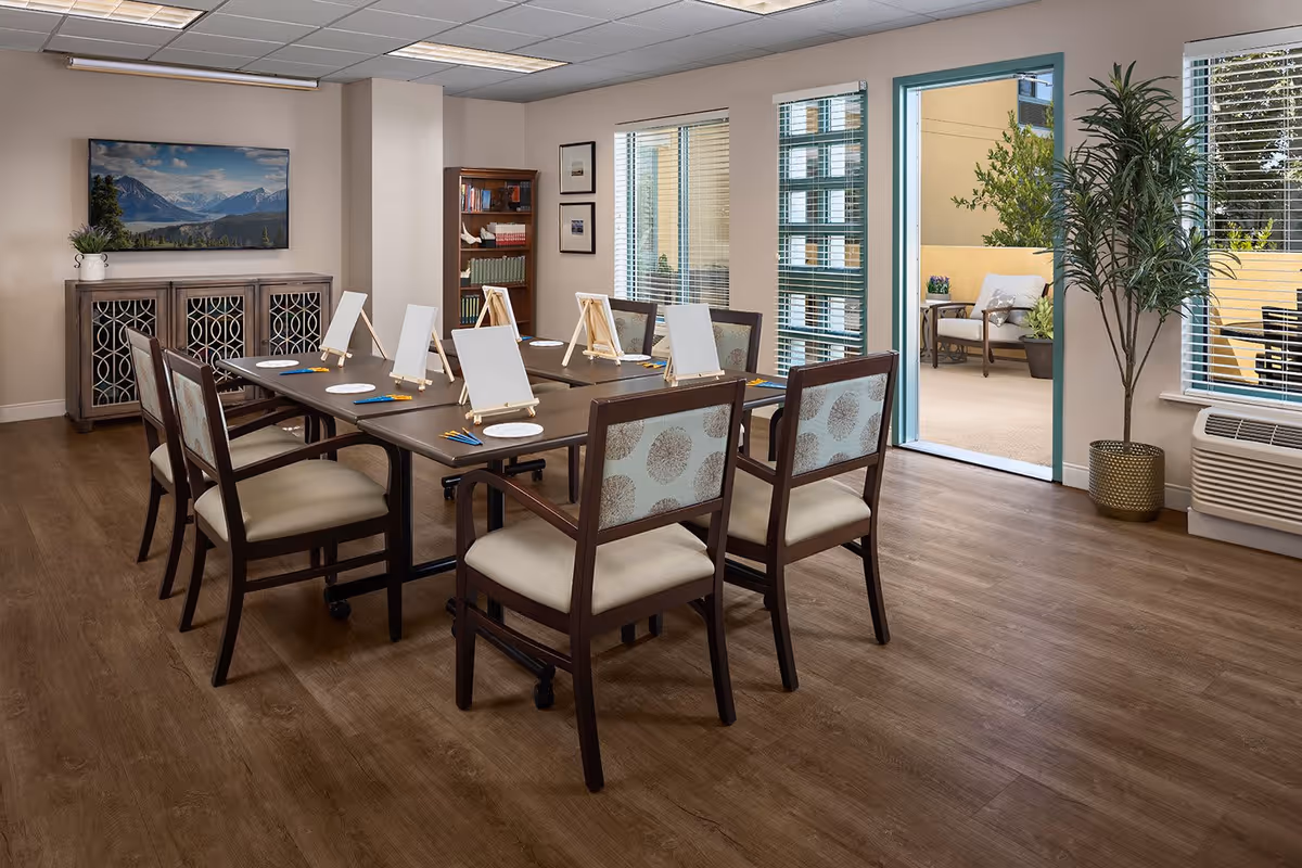 A well-lit activity room with a rectangular table surrounded by eight chairs, each with a small easel, paintbrushes, and paper plates for painting. The room has wooden flooring, a cabinet with a landscape painting above it, a bookshelf, large windows with blinds, and a door leading to an outdoor patio with seating and plants.