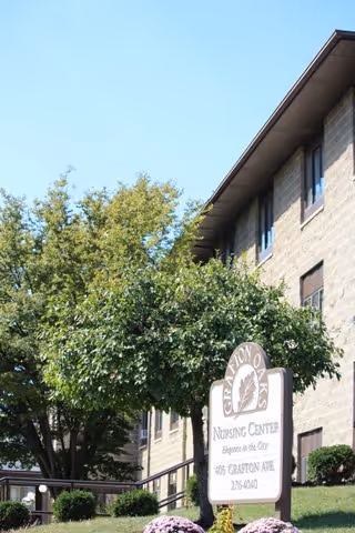 Exterior view of a multi-story brick building with several windows, partially obscured by green trees and bushes. In front of the building is a sign that reads 'Grafton Oaks Nursing Center Elegant in the City 465 Grafton Ave 276-4040'. The sky is clear and blue.