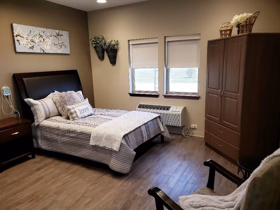 A cozy bedroom with a neatly made bed featuring striped bedding and decorative pillows, including one with the word 'love'. The room has two windows with white blinds, a wooden nightstand with a drawer, a wooden wardrobe with baskets and flowers on top, and a comfortable chair with a light-colored throw. The walls are beige, and there is a floral painting and two wall-mounted planters with greenery.