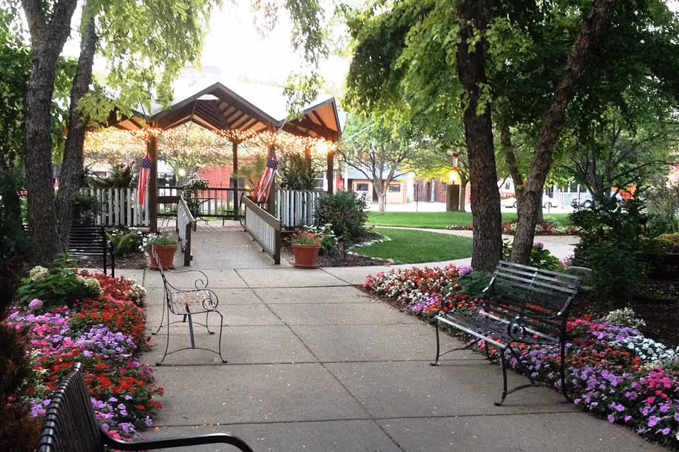 A peaceful outdoor garden area with a paved walkway, colorful flower beds on both sides, several benches, and a wooden gazebo decorated with string lights and American flags. Trees provide shade and greenery surrounds the space.