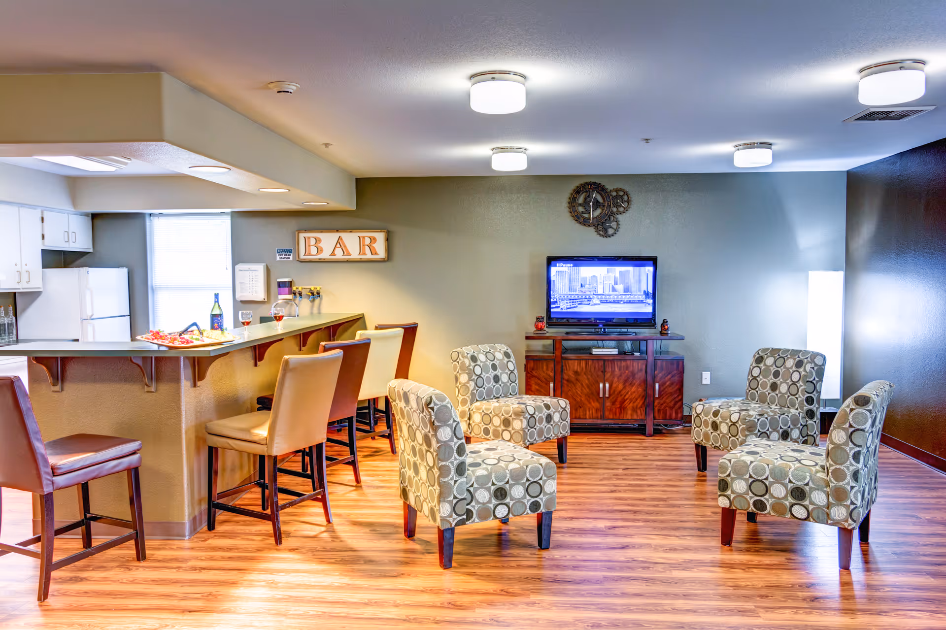 A cozy common area in an assisted living facility featuring a small bar with four bar stools, a kitchen area with white cabinets and a refrigerator, and a seating area with four patterned armchairs arranged around a TV on a wooden stand. The room has wood flooring and ceiling lights, with a decorative wall clock above the TV and a sign that reads 'BAR' above the bar counter.