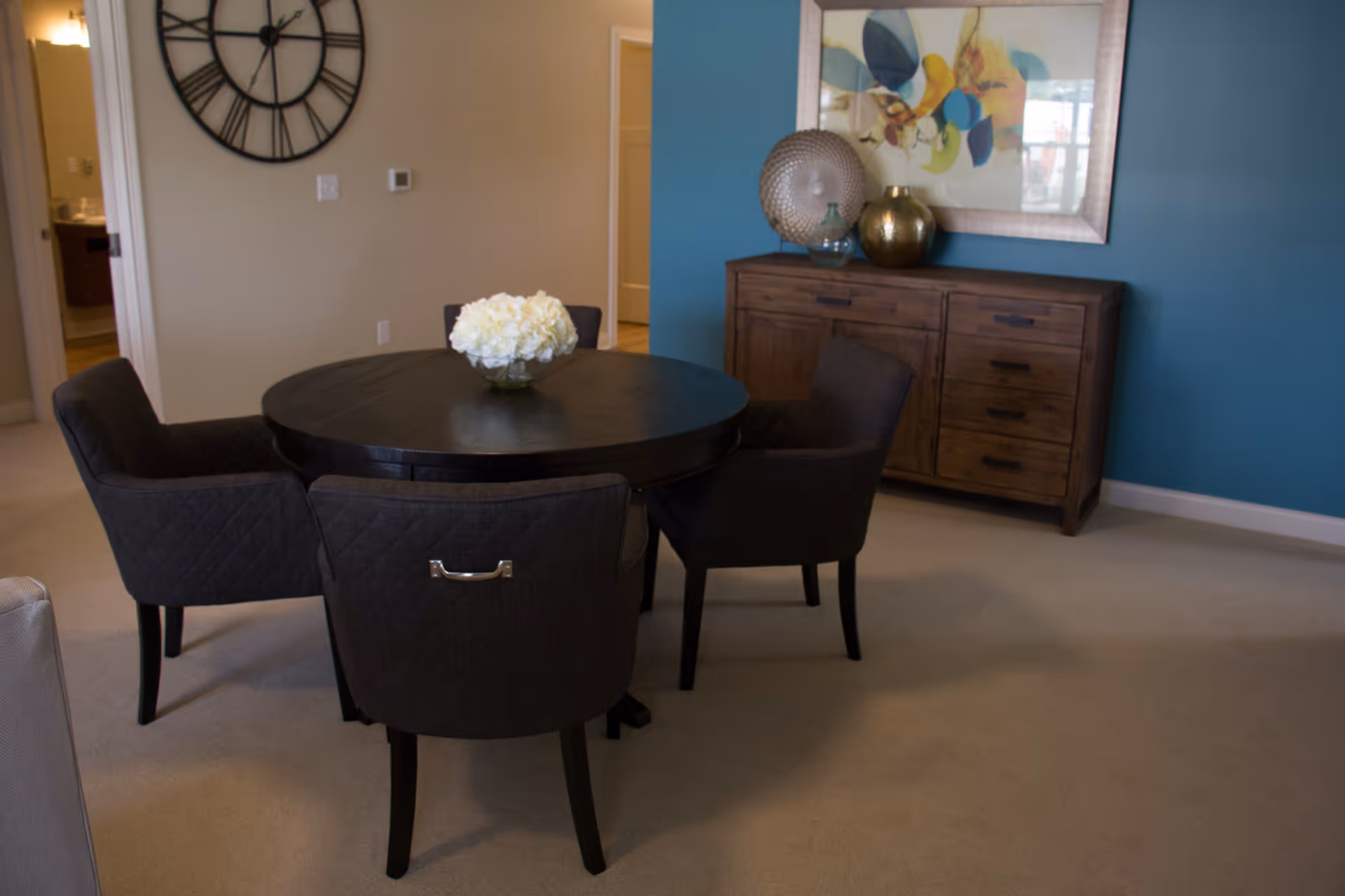 A dining area with a round dark wooden table surrounded by four dark upholstered chairs. A vase with white flowers is placed in the center of the table. In the background, there is a wooden sideboard with decorative items and a framed abstract floral artwork hanging on a blue accent wall. A large black wall clock is mounted on a beige wall near an open doorway.
