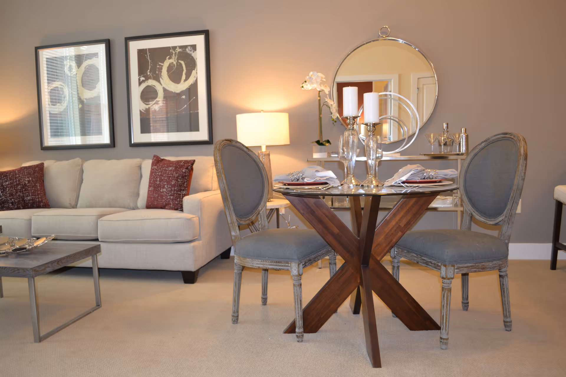 A cozy living room area featuring a beige sofa with two red patterned pillows, a glass-top round dining table with a wooden X-shaped base, and two upholstered chairs. The table is set with plates, napkins, and glass candle holders. Behind the table is a console table with decorative items including a round mirror, candles, and a flower vase. Two abstract framed artworks hang on the wall above the sofa, and a table lamp provides warm lighting.