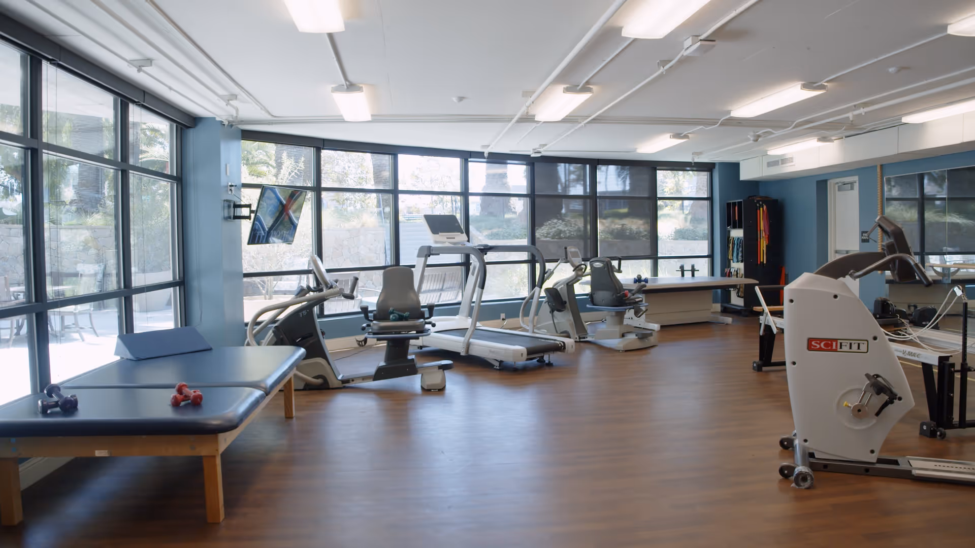 A bright and spacious exercise room with large windows letting in natural light. The room contains various fitness equipment including treadmills, recumbent bikes, a rowing machine, and a padded exercise table with small dumbbells on it. The floor is wooden, and the walls are painted blue.
