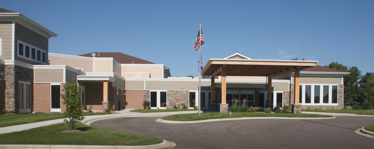 Front exterior view of Oak Park Place Janesville facility showing a modern building with a covered entrance supported by wooden beams, an American flag and another flag on flagpoles, surrounded by well-maintained grass and a paved driveway.