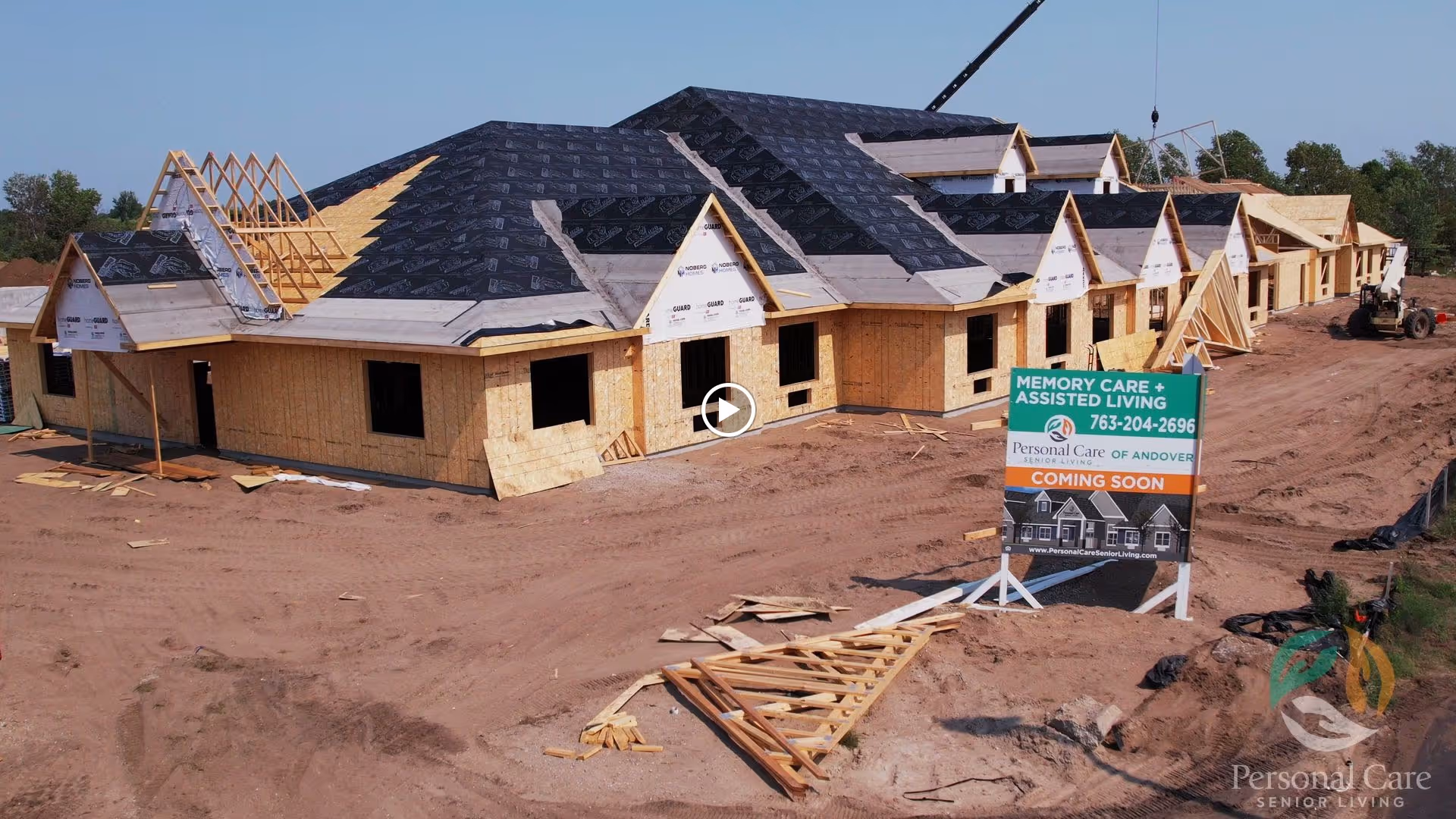A large building under construction with wooden framing and roofing materials partially installed. There is a sign in front of the construction site that reads 'Memory Care + Assisted Living, Personal Care of Andover, Coming Soon' along with a phone number and website. The ground around the building is dirt with construction materials scattered.