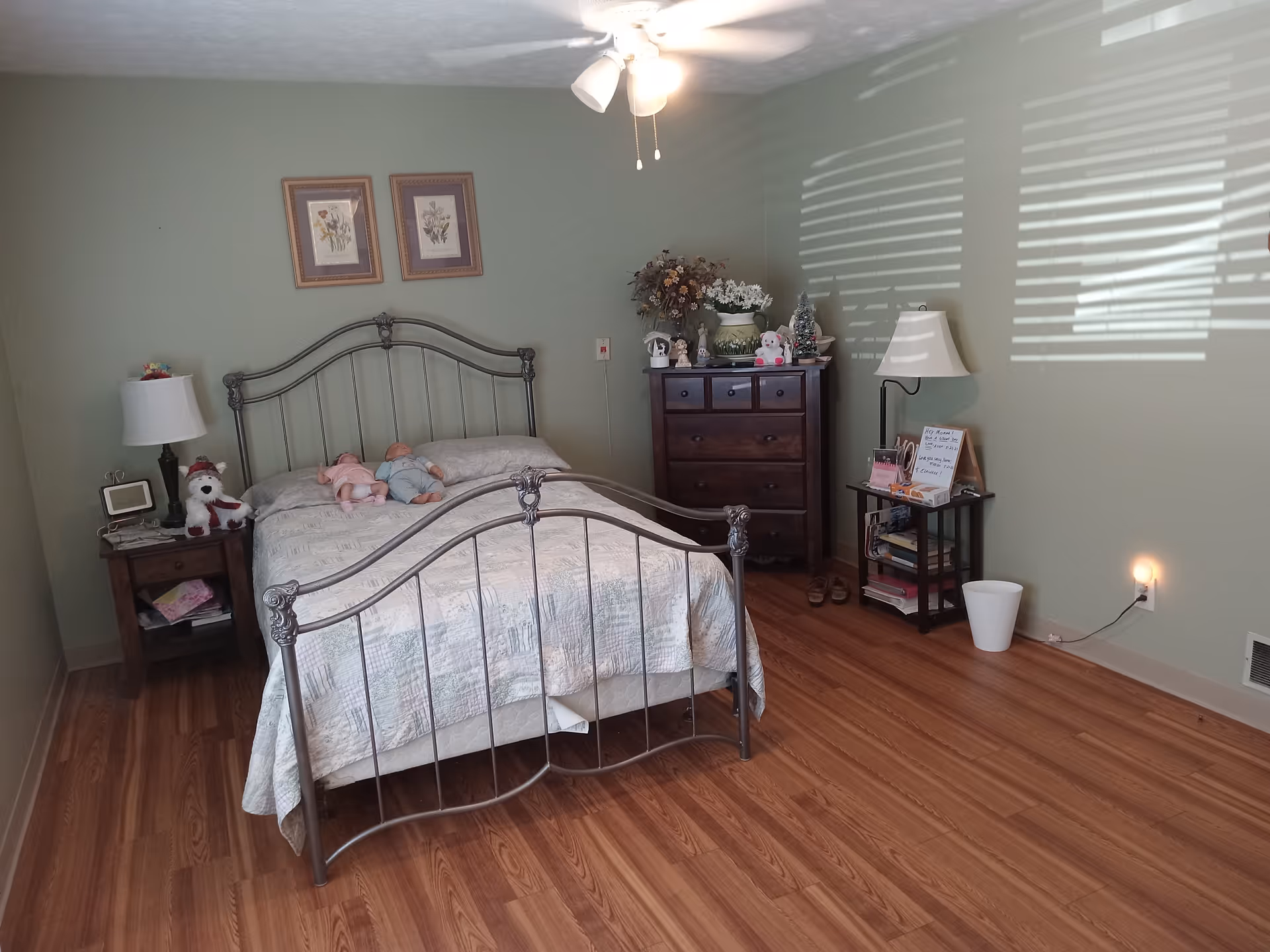 A bedroom with a metal bed frame and a quilted bedspread. Two baby dolls are placed on the bed. There is a wooden nightstand with a lamp and a stuffed dog toy on the left side of the bed. On the right side, there is a wooden dresser with decorative items including flowers and small figurines. A small table with a lamp, books, and a whiteboard is next to the dresser. The room has light green walls, wooden flooring, and sunlight casting shadows through window blinds.