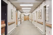 A long, well-lit hallway in a skilled nursing and rehabilitation center with beige walls, carpeted floor, handrails on both sides, framed pictures on the walls, and doors leading to rooms on either side.