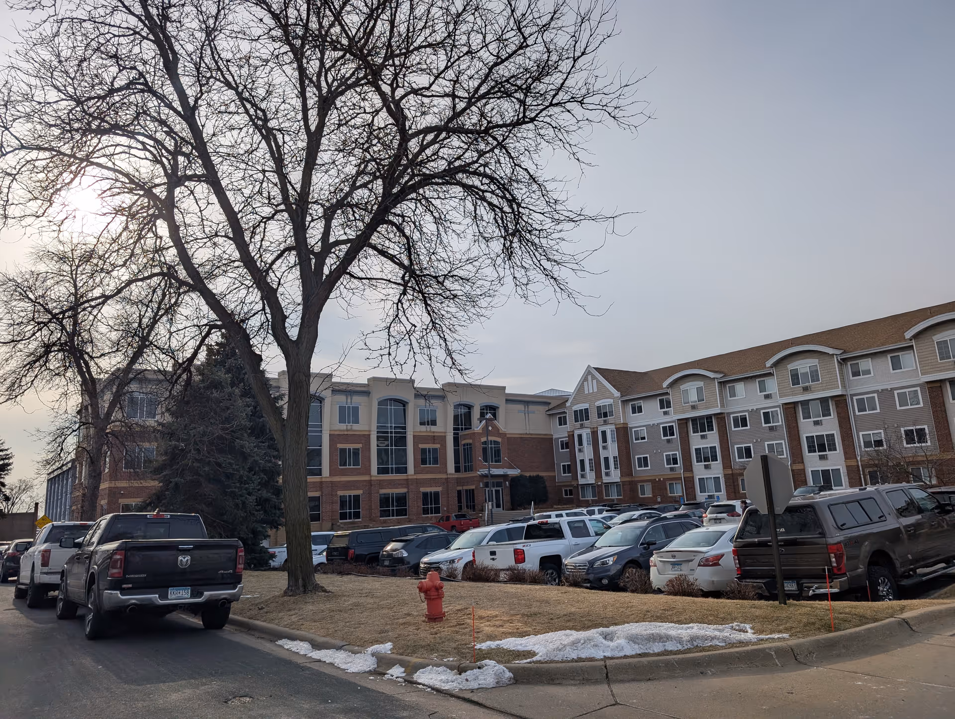 Exterior view of a multi-story senior living facility named Mainstreet Village with a parking lot filled with cars in front. Leafless trees and patches of snow are visible on the grass near the sidewalk.