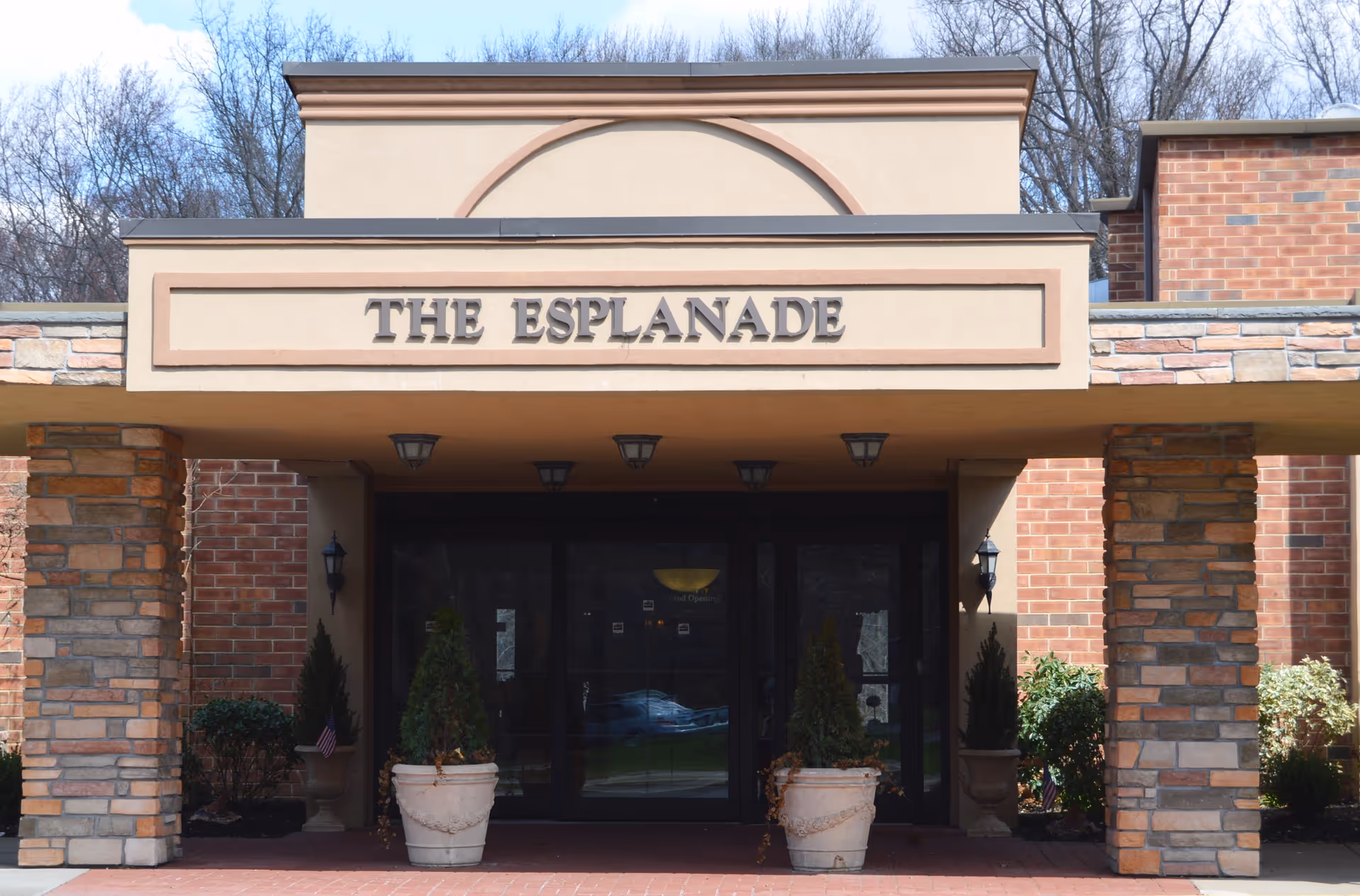 Entrance of The Esplanade building with stone pillars, two large potted plants, and glass double doors under a beige overhang with the name THE ESPLANADE displayed above.