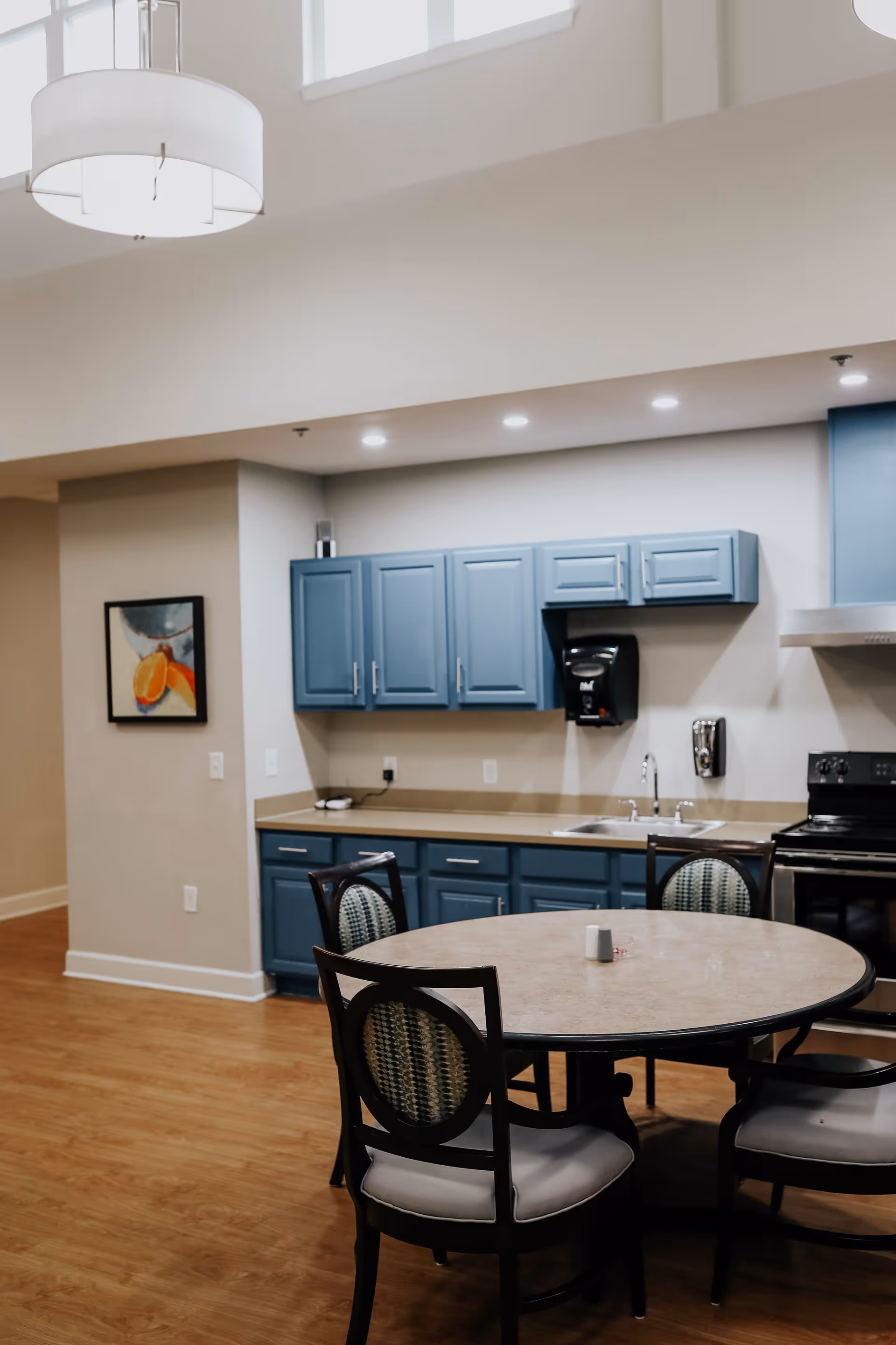 Interior view of a kitchen area with blue cabinets, a round dining table with four chairs, a sink, stove, and a wall-mounted soap dispenser. The room has wooden flooring and a modern light fixture hanging from the ceiling.