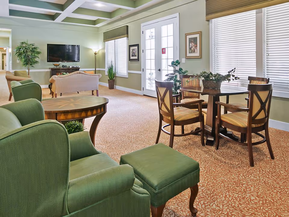 A bright and spacious living room area in an assisted living facility featuring green upholstered armchairs with matching ottomans, a round wooden table with four cushioned chairs, potted plants, a wall-mounted flat screen TV, and large windows with blinds allowing natural light to fill the room.