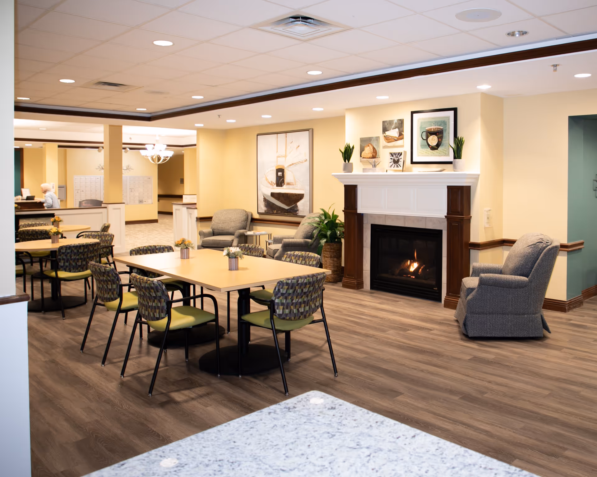 A cozy common area in a senior living facility featuring a fireplace with a fire burning, several armchairs, and tables with chairs arranged for socializing or dining. The walls are painted light yellow, decorated with framed artwork and plants. The flooring is wood-style laminate, and the ceiling has recessed lighting.