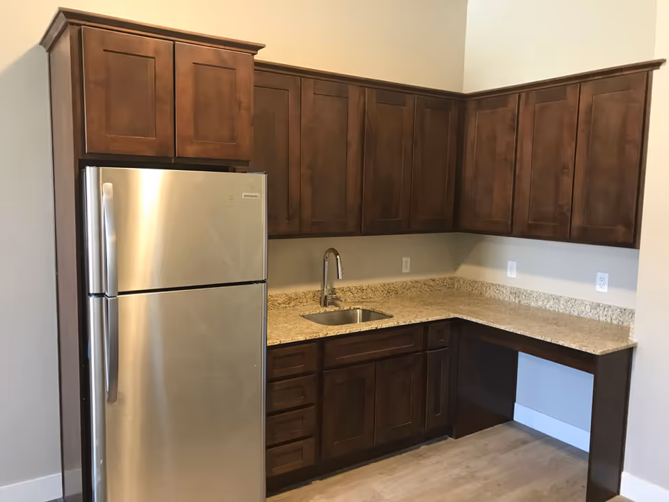 Compact kitchen with stainless steel refrigerator, granite countertops, sink, and dark wood cabinets.