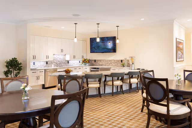 A bright and spacious kitchen and dining area with white cabinets, a stone backsplash, and a large island with four cushioned bar stools. There are two round dining tables with wooden chairs featuring cushioned seats and backs. The floor is carpeted with a checkered pattern, and a flat-screen TV is mounted on the wall above the kitchen counter. Pendant lights hang above the island, and there are small flower arrangements on the dining tables.