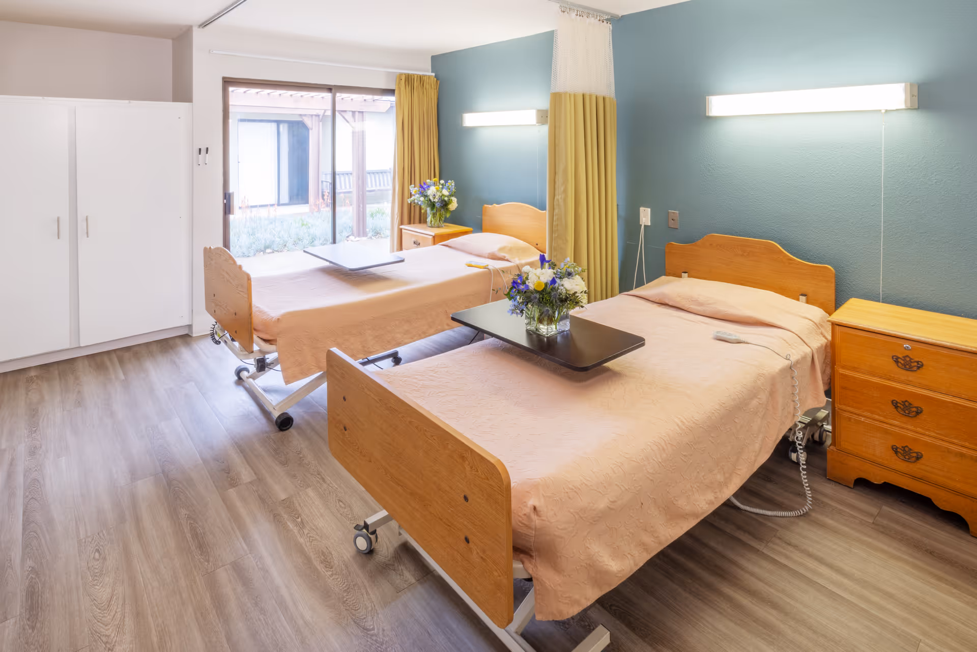 A bright and clean room with two hospital-style beds, each with a light pink bedspread and a small table with a vase of flowers. The room has light wood flooring, a large window with yellow curtains, a white wardrobe, and a blue accent wall with two wall-mounted lights above the beds.