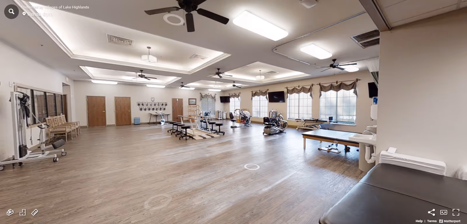 A spacious rehab gym with wooden flooring, multiple exercise machines, therapy tables, chairs, and large windows with valances allowing natural light. The ceiling has recessed lighting and ceiling fans, and there are several doors along the far wall.